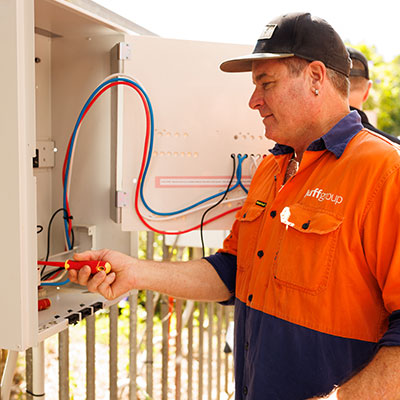 An electrician in an orange shirt and vest is focused on an electrical panel, representing Huffgroup electrical