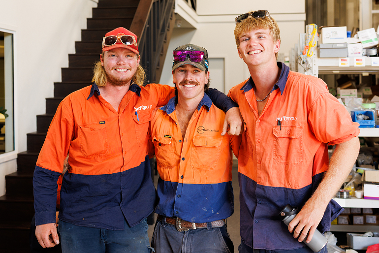 Three men in orange and blue work shirts standing together, representing Huffgroup Electrical Services