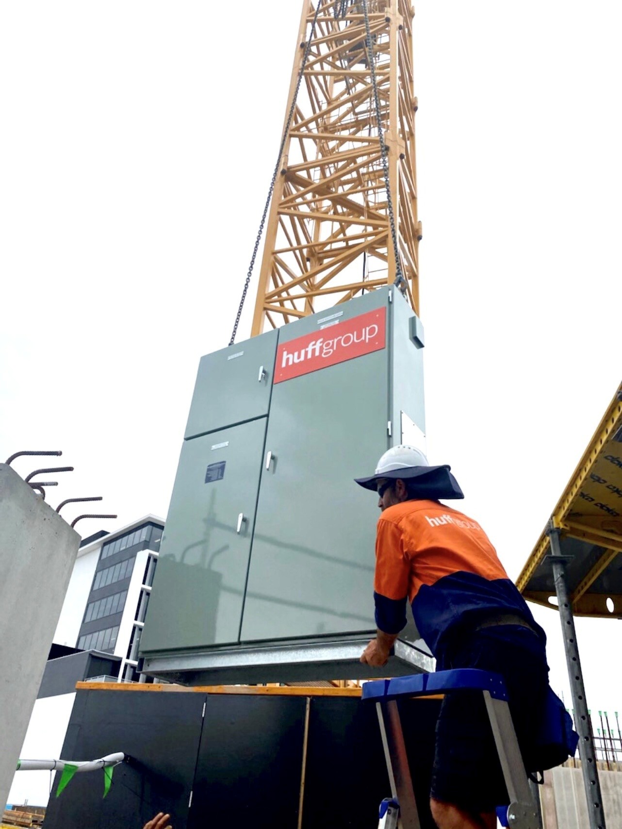 A man operates a crane in front of a building, with the Huff Group logo visible on the crane